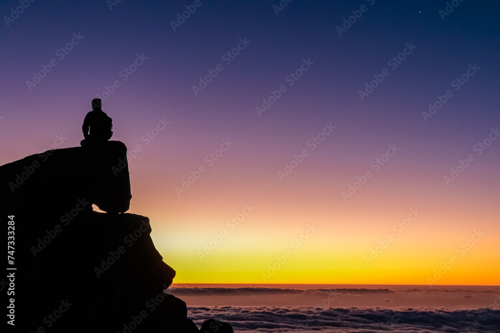 Silhouette of person sitting on massive rock watching romantic sunrise on mountain peak Pico do Areeiro, Madeira island, Portugal, Europe. Panoramic view of clouds over Atlantic Ocean. Orange red sky