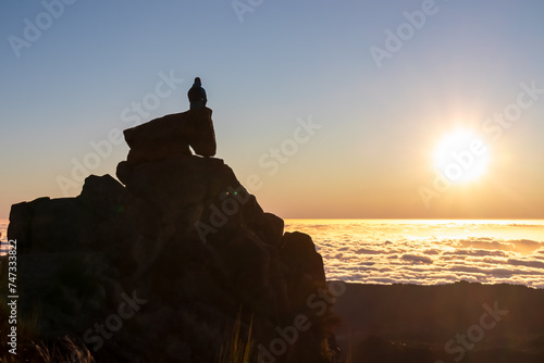 Silhouette of person sitting on massive rock watching romantic sunrise on mountain peak Pico do Areeiro, Madeira island, Portugal, Europe. Panoramic view of clouds over Atlantic Ocean. Orange red sky