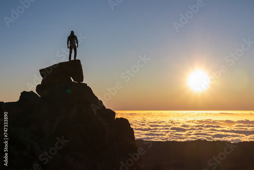 Silhouette of person standing on massive rock watching romantic sunrise on mountain peak Pico do Areeiro, Madeira island, Portugal, Europe. Panoramic view of clouds over Atlantic Ocean. Orange red sky