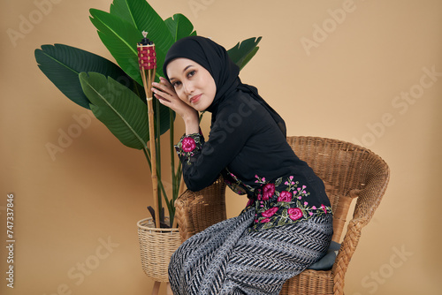 Beautiful Asian model wearing black kebaya dress with hijab, sitting on a rattan chair with bamboo torch and artificial plant as background. Eidul fitri festival, fashion and beauty concept. photo