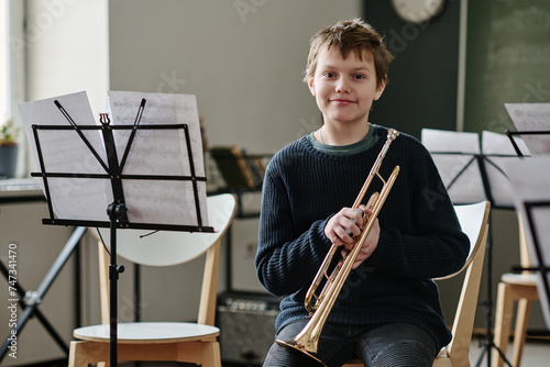 Medium portrait of cheerful Caucasian teen boy holding trumpet sitting in classroom equipped with music stands