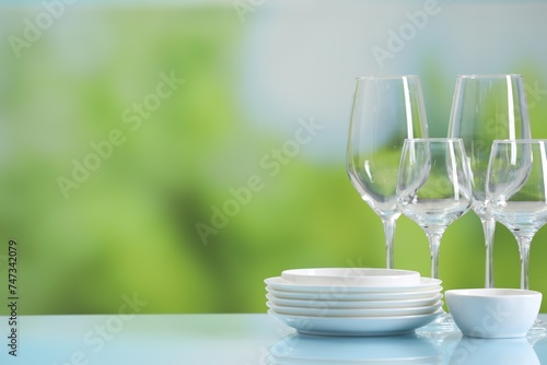 Set of many clean dishware and glasses on light blue table against blurred green background. Space for text