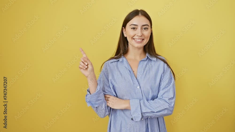 Smiling young hispanic woman, beautiful and confident, happily pointing to the side with her hand and finger. wearing a shirt, she stands isolated against a yellow background. Smiling young hispanic woman, beautiful and confident, happily pointing to the side with her hand and finger. wearing a shirt, she stands isolated against a yellow background.