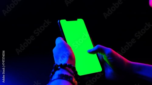 Mobile phone in hand. Holding smartphone with red blue neon lighting on dark background. Green chromakey screen with markers. Hand lifts phone up and holds it still.