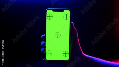 Mobile phone in hand. Holding smartphone with red blue neon lighting on dark background. Green chromakey screen with markers. Hand lifts phone up and holds it still.
