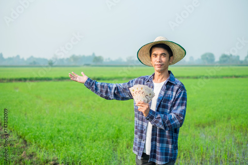 Asian male farmer dressed in blue jeans Stand and pose holding banknote money at a green rice field.