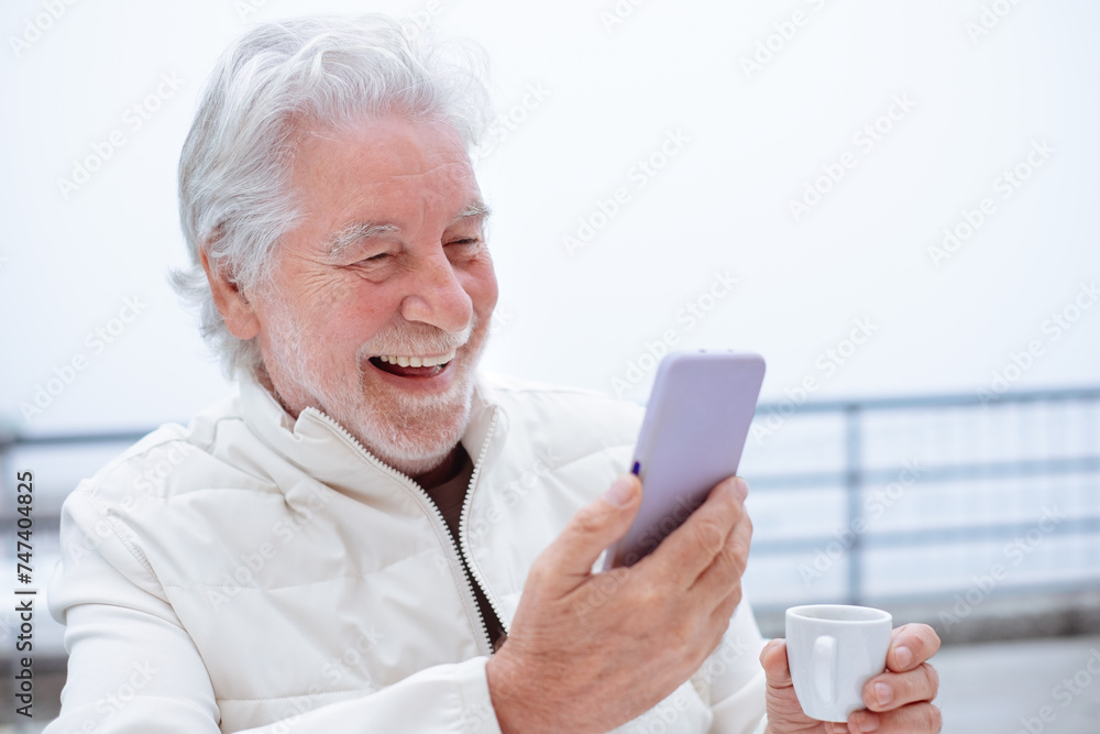 Video call concept. Portrait of happy handsome white haired senior man holding a coffee cup talking by mobile phone with online webcam connection Video call concept. Portrait of happy handsome white haired senior man holding a coffee cup talking by mobile phone with online webcam connection