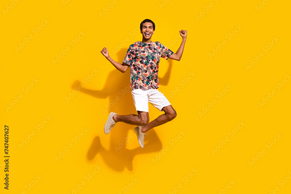Full length photo of funky lucky guy dressed print t-shirt jumping high rising fists isolated yellow color background