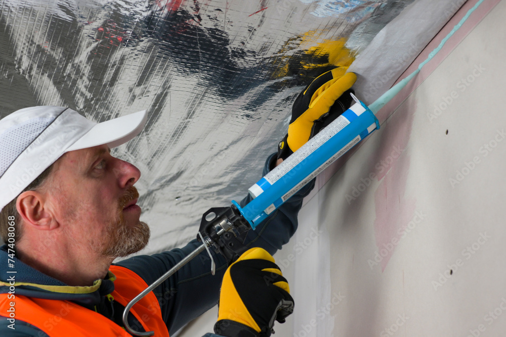 Fototapeta premium a worker in a vest, cap and gloves uses a vapor barrier glue gun