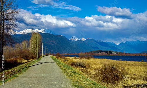 Spring sunny day in the Pitt Valley, the road along the river, green  grass on the sidelines, bare trees against a mountain range and a blue cloudy sky