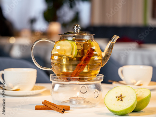 Canvas Print Herbal tea with apple and cinnamon in transparent glass teapot with warmer and candle on table in restaurant