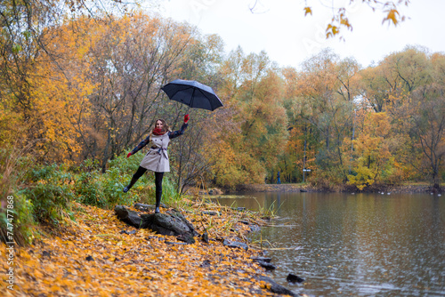 A girl in an autumn park stands on a stone and pretends to fly away on an umbrella. Portrays Mary Poppins. On the background there is a multicolored autumn forest of yellow  orange  red colors