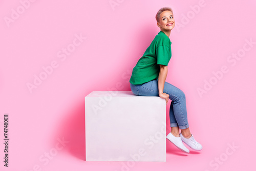 Full length photo of cheerful woman wear t-shirt sit on platform dreamy look at offer empty space isolated on pink color background