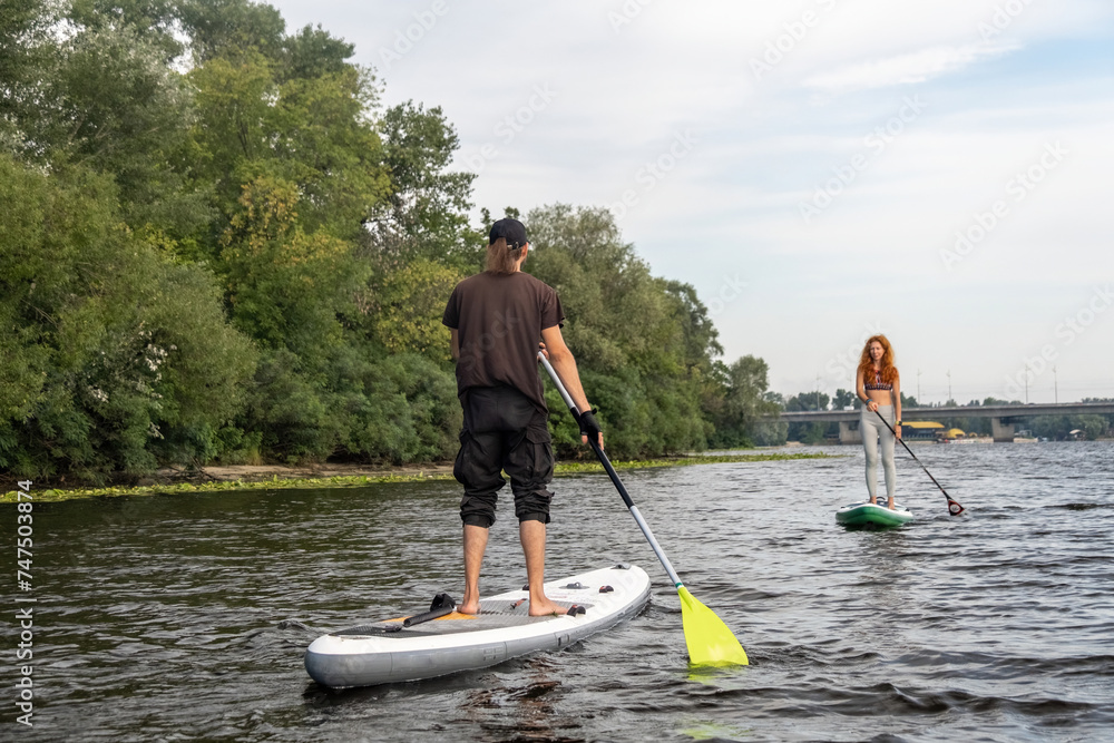Man and woman balance on paddleboards having active rest on weekend. Friends enjoy training session in natural surrounding