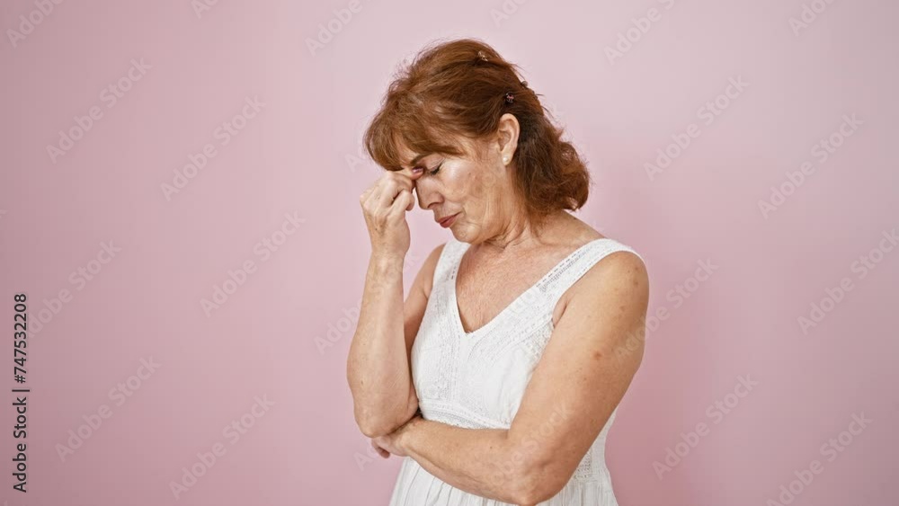 Exhausted middle-age woman in dress touches fatigued eyes, rubbing pained nose. stress-induced headache evident. standing in frustration against a solo pink backdrop. Exhausted middle-age woman in dress touches fatigued eyes, rubbing pained nose. stress-induced headache evident. standing in frustration against a solo pink backdrop.