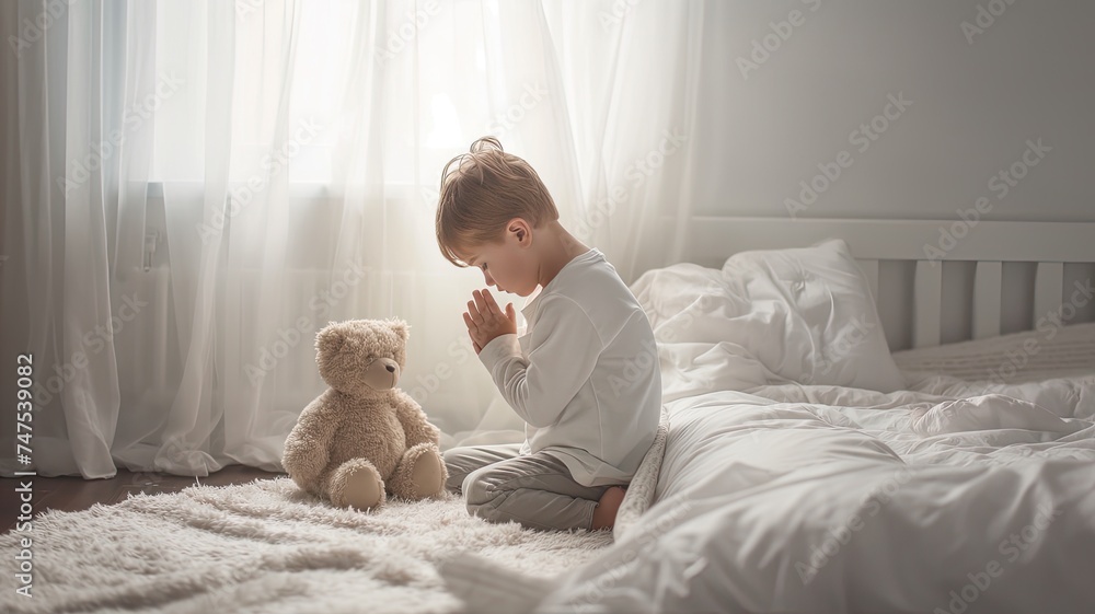 a three-year-old boy kneeling in prayer beside his bed in his bedroom, head bowed, with a teddy bear nearby, against a backdrop of soft white pastel colors. a three-year-old boy kneeling in prayer beside his bed in his bedroom, head bowed, with a teddy bear nearby, against a backdrop of soft white pastel colors.