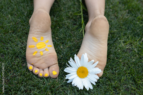 bare feet of a child lying on green grass . A daisy flower between the toes, a painted yellow sun on one foot. cheerful positive atmosphere, happy childhood. Hello summer