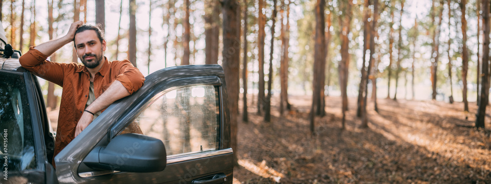 A young man stands next to a car with an upper tourist trunk in a pine forest on the shore of a lake at sunset. A young man stands next to a car with an upper tourist trunk in a pine forest on the shore of a lake at sunset.