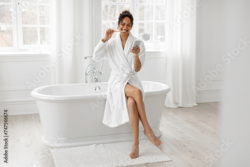 Young black woman in bathrobe using smartphone and brushing teeth, sitting on bathtub, holding phone, full length, free space