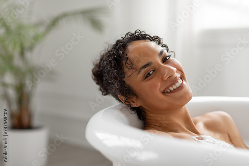 Relaxed black woman enjoying foamy hot bath, leaning on tub and smiling at camera, relaxing at weekend at home, copy space photo