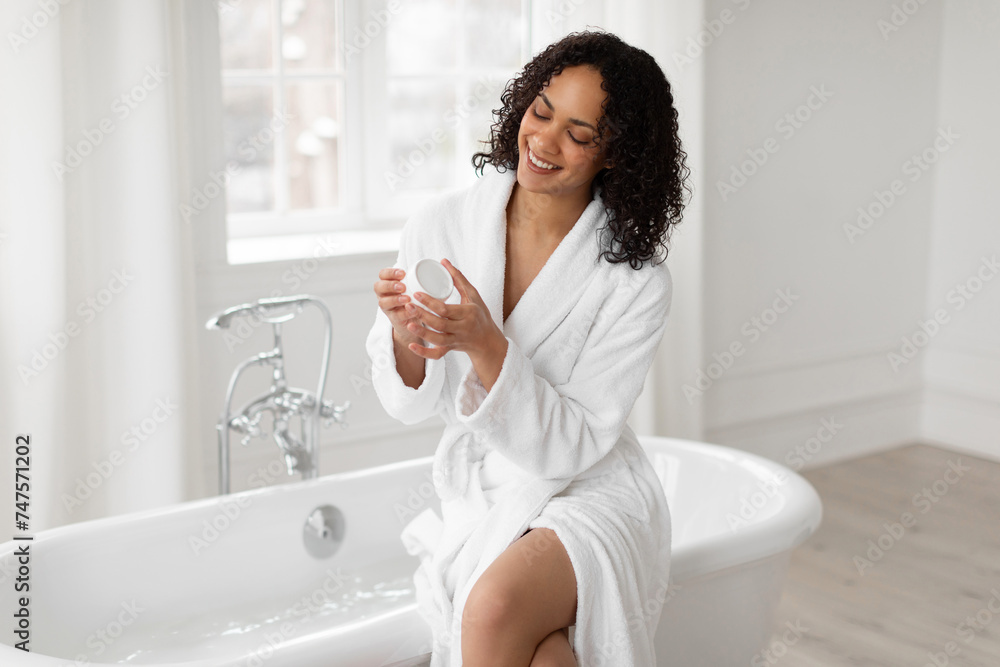 Skincare concept. Happy black lady holding cream moisturizer, doing female beauty rituals, recommending skin care cosmetic products, bathroom interior