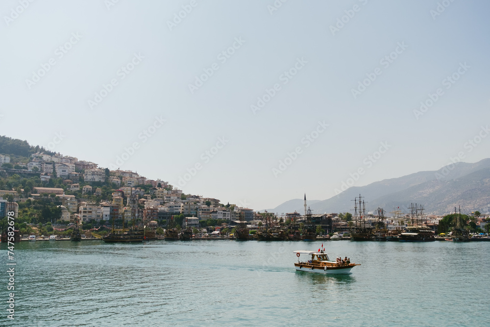 A small boat sails across calm waters with a bustling hillside town and ships in the background