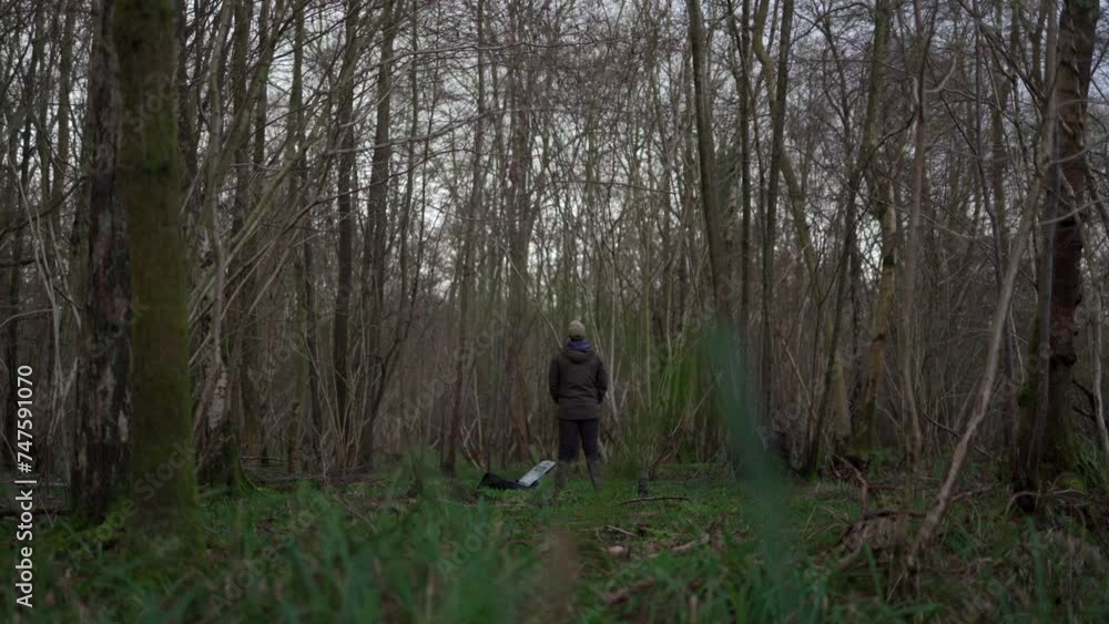 A man standing in a dull woodland looking up at the tree canopy beside his open backpack A man standing in a dull woodland looking up at the tree canopy beside his open backpack