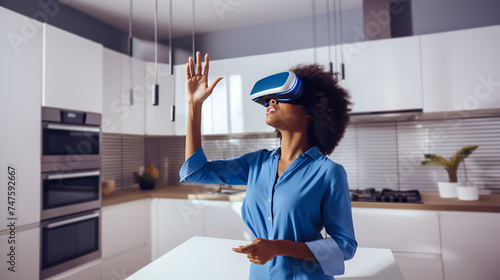 A black woman wearing augmented virtual reality glasses and rich blue clothes gesticulates with her hands while controlling a virtual screen while standing in a modern home kitchen.