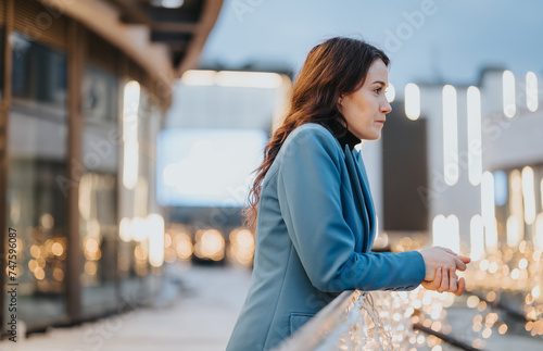 A pensive woman in a blue blazer stands outdoors at evening, surrounded by warm city lights, embodying urban contemplation and serenity.