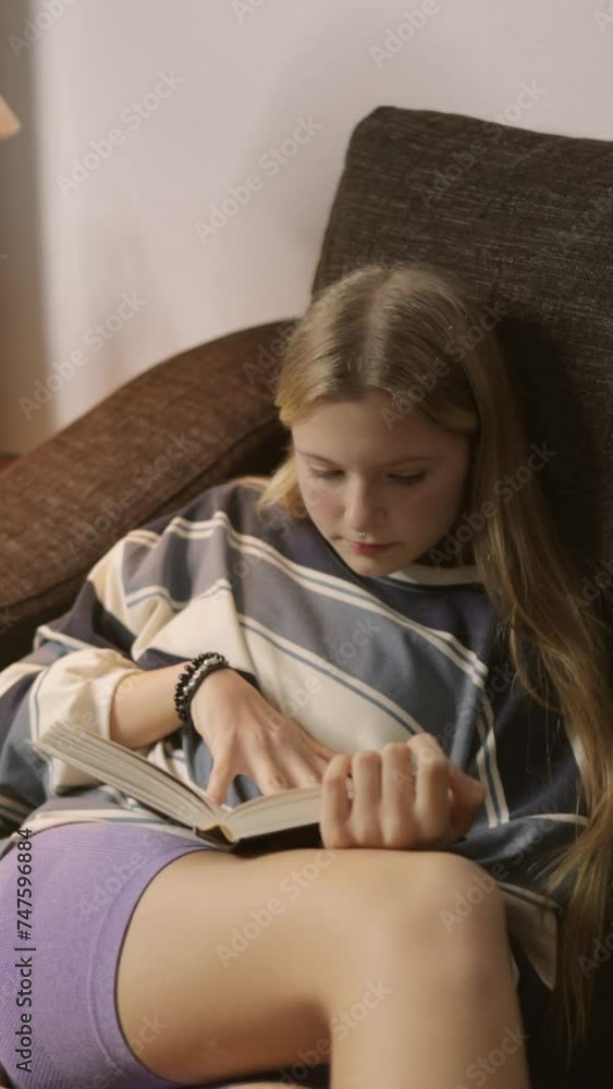 A teenage girl sits comfortably on the couch, her attention fully absorbed in the pages of a book she holds in her hands. The tranquil atmosphere of the room provides the perfect backdrop.Vertical A teenage girl sits comfortably on the couch, her attention fully absorbed in the pages of a book she holds in her hands. The tranquil atmosphere of the room provides the perfect backdrop.Vertical