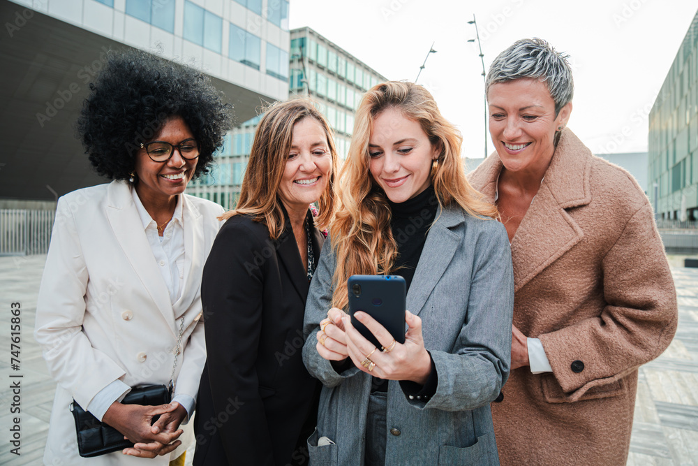 Fototapeta premium Group of mid adult business women using a cellphone to work. Successful corporate female coworkers watching a smartphone. Professional office ladies sharing with a mobile phone on a executive meeting