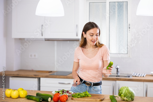 Portrait of young joyful woman standing in home kitchen and chopping green pepper, preparing vegetable dish