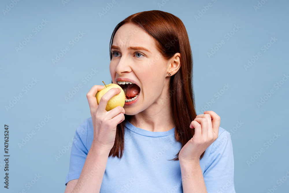 Red haired smiling woman with braces in blue shirt holding green apple and biting isolated Red haired smiling woman with braces in blue shirt holding green apple and biting isolated