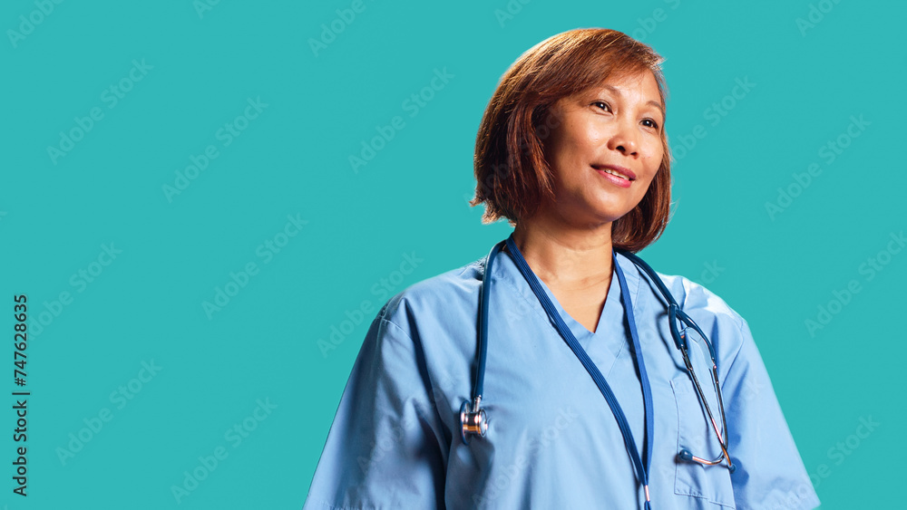 Happy proficient BIPOC nurse smiling while looking at camera. Portrait of cheerful asian healthcare specialist wearing professional scrubs, isolated over blue studio background Happy proficient BIPOC nurse smiling while looking at camera. Portrait of cheerful asian healthcare specialist wearing professional scrubs, isolated over blue studio background