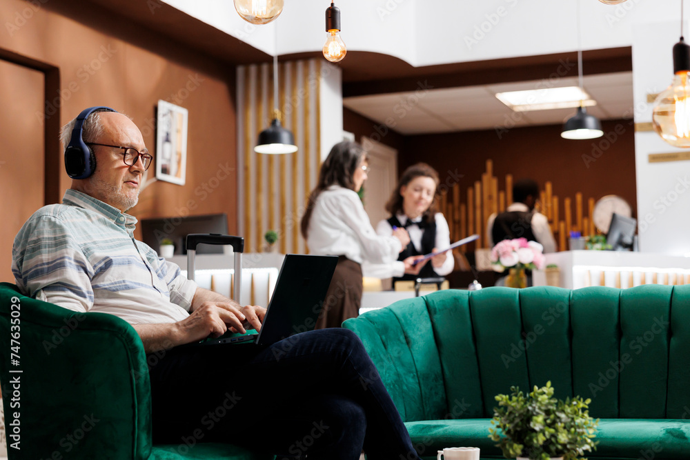 Caucasian old man using wireless computer while elderly female tourist checks in at front desk. Retired senior male traveler sitting in lounge area surfing the net on laptop and wearing headphones. Caucasian old man using wireless computer while elderly female tourist checks in at front desk. Retired senior male traveler sitting in lounge area surfing the net on laptop and wearing headphones.