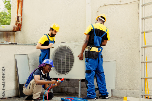 Diverse team of seasoned wiremen installing new external air conditioner for customer. Licensed servicemen tasked to optimize new HVAC system's performance, ensuring it runs at perfect efficiency photo