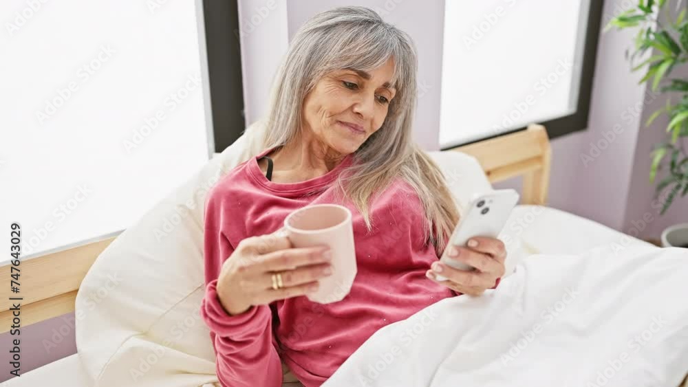 A mature hispanic woman with grey hair relaxes in bed at home, holding a coffee mug and smartphone, portraying leisure in a bedroom.