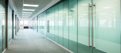 The long hallway is lined with transparent glass walls and doors, allowing natural light to illuminate the space. The sleek design gives a modern and professional atmosphere to the office environment.