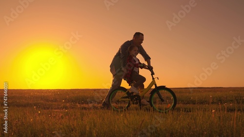 Dad helps his daughter ride bike. Kid Girl learns to ride bike. Happy family nature. Father teaching kid daughter to ride bike in park. Child rides bike. Child, dad play together, sun. Childhood dream