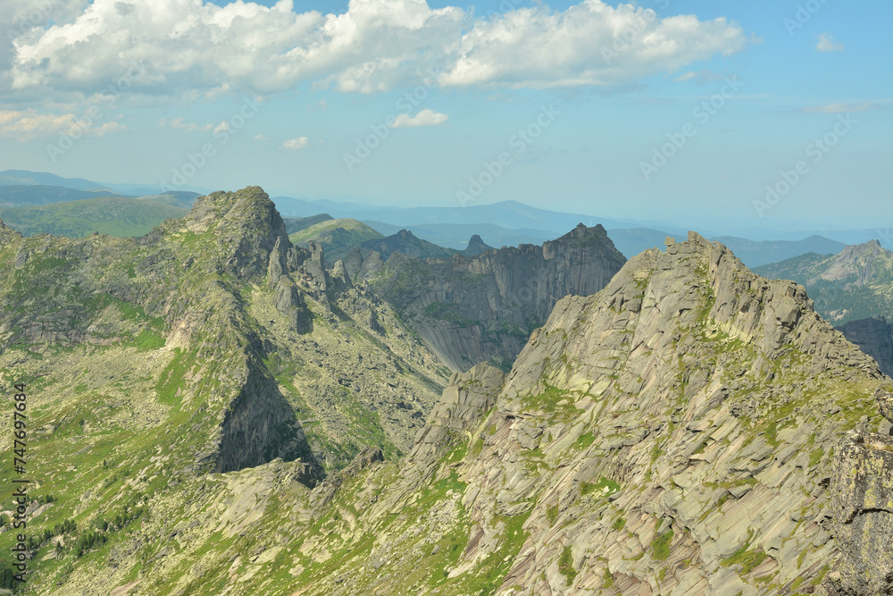 A chain of tall, jagged rocks curves around the mountain ridges under a cloudy summer sky.