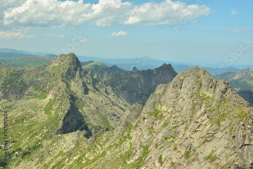 A chain of tall, jagged rocks curves around the mountain ridges under a cloudy summer sky.