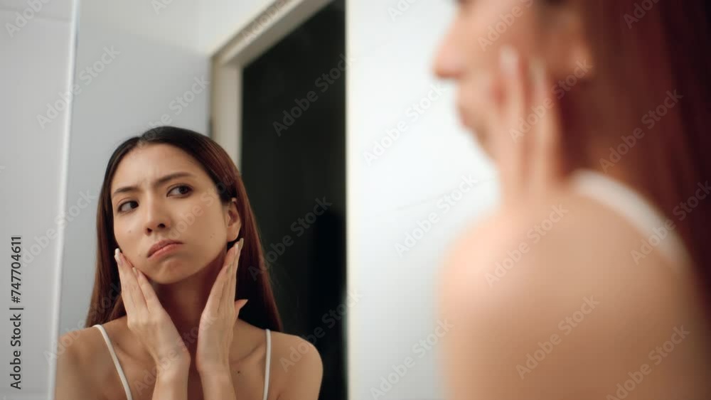 Worry feeling of asian woman touching unhealthy face skin looking at mirror and checking dry irritated face skin in bathroom. Unhappy Young female looking at red acne spots on face skin care. Worry feeling of asian woman touching unhealthy face skin looking at mirror and checking dry irritated face skin in bathroom. Unhappy Young female looking at red acne spots on face skin care.