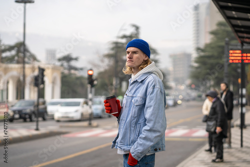 Long-haired hipster guy in stylish outfit, standing in city street with cup of coffee, looking into distance, waiting for taxi, friend. Carefree fashionable male, representative of generation z