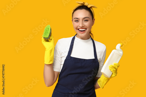 Portrait of happy young housewife in apron with cleaning supplies on yellow background photo