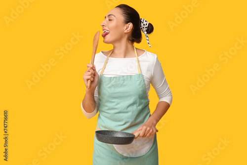 Portrait of beautiful young housewife in apron with frying pan and wooden spatula on yellow background