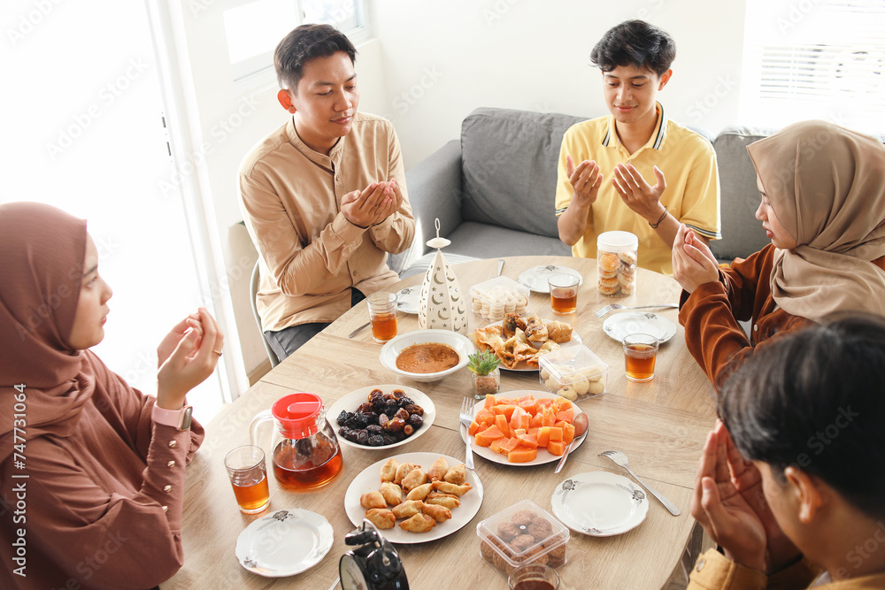 Group of young Asian muslim man and woman raising hands praying before eating together. Breakfasting, religious people and Islamic tradition concept
