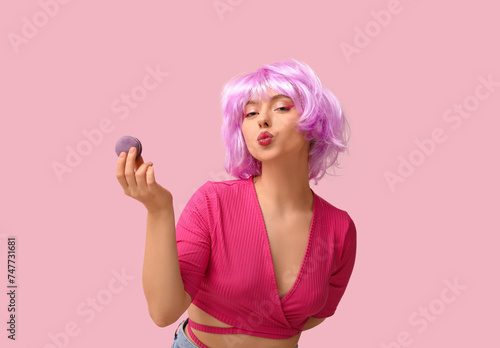 Beautiful young woman in wig with purple sweet macaroon on pink background