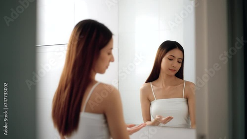 Happy attractive asian woman applying lotion cream or moisturizer skin care on hand and body in front of mirror in bathroom. Pretty female applying cream her beauty smooth body skin care treatment. photo