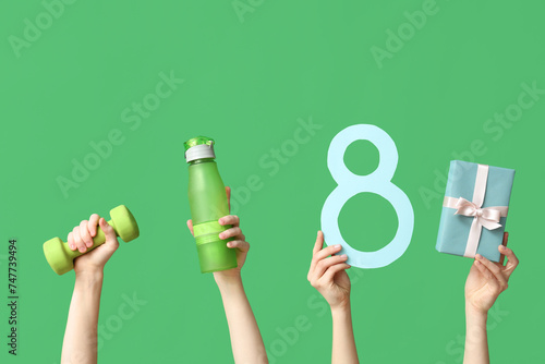 Female hands with paper figure 8, sports equipment and gift box for International Women's Day on green background