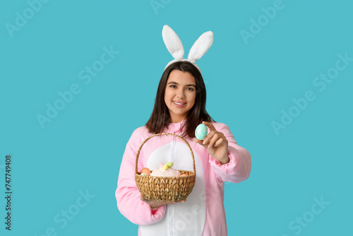 Beautiful young woman in bunny costume holding wicker basket with Easter cake and eggs on blue background